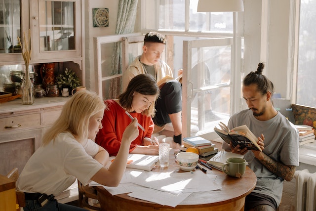 Four student roommates sit in a brightly kit kitchen and tudy around a kitchen table littered with papers and books